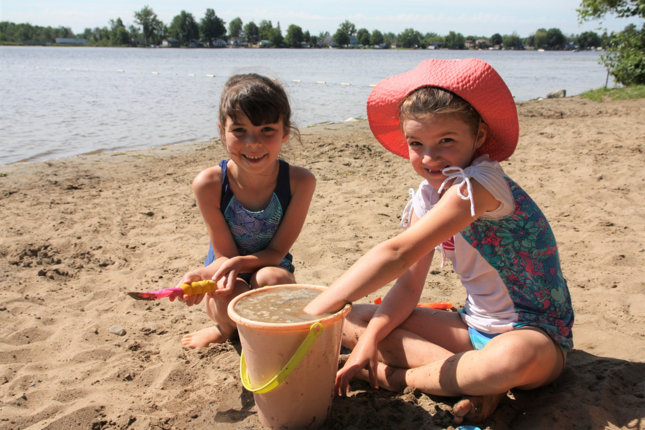 Two young girls smile and play with buckets in the sand in front of the Rideau River.