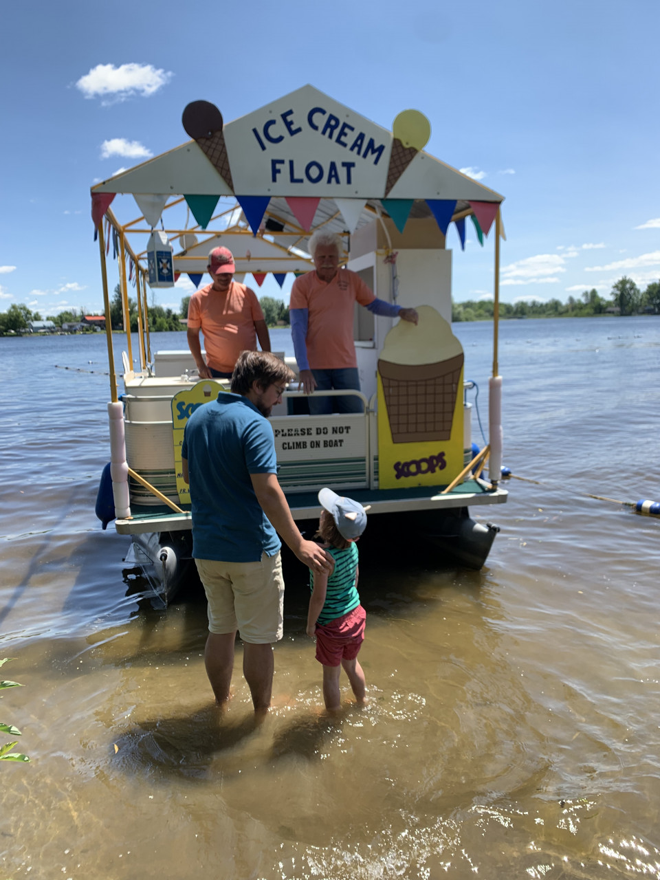 A man and child stand in front of a pontoon boat decorated with ice cream cone signs in the shallow waters at Baxter Beach.