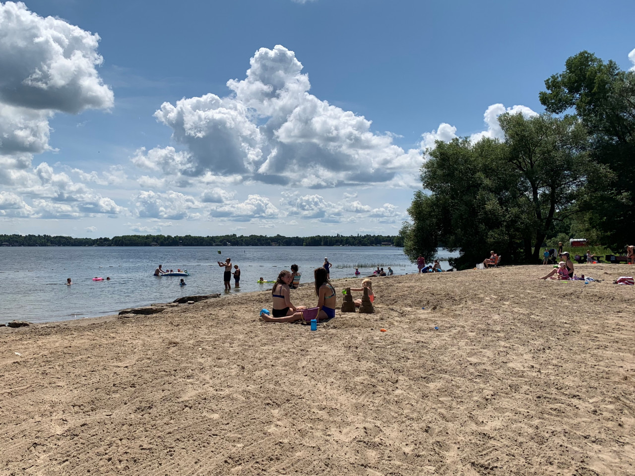 People play in the sand in front of the blue waters of Lower Rideau Lake. White puffy clouds dot the blue sky.