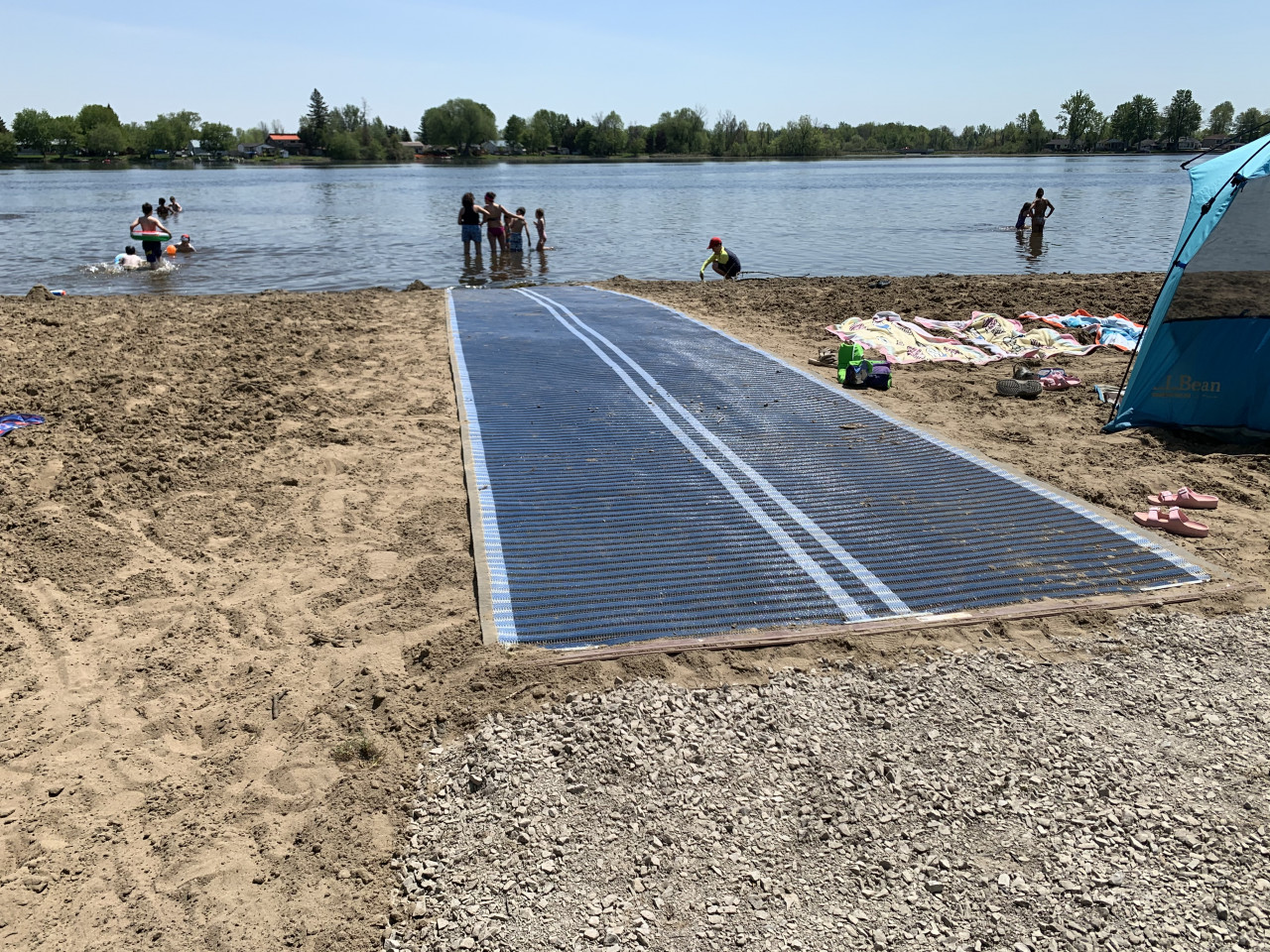 An accessible mat runs across a sandy beach down to the Rideau River while beach-goes play in the water.