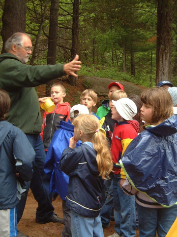 A teacher gestures to students who have gathered in the forest for outdoor education